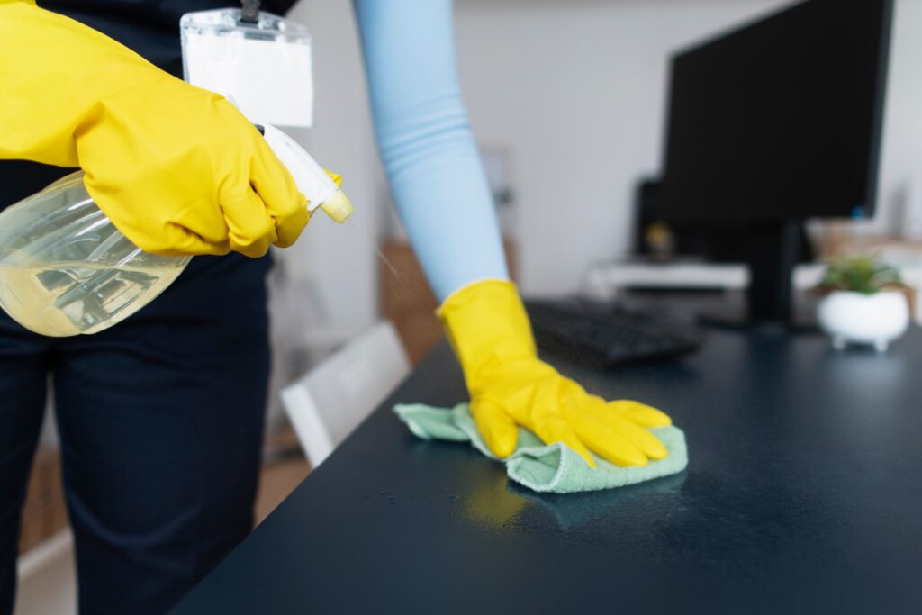 Person in yellow gloves applying green cloth to dark desk surface near computer monitor and keyboard workspace sanitization