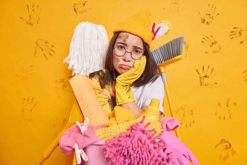 Overwhelmed woman in yellow gloves and cap holding cleaning products against muddy handprint wall expressing cleaning frustration